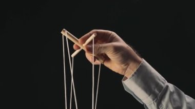 The puppeteers hand controls the puppet with a wooden manipulator and strings. The marionettist controls and pulls the strings on a black background. The concept of dependence, dominance, managing