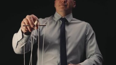 A businessman in a white shirt and black tie controls a puppet with a wooden manipulator and strings. A puppeteer controls a doll marionette on a black isolated background. The concept of control