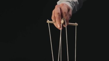 The puppeteers hand controls the puppet with a wooden manipulator and strings. The marionettist controls and pulls the strings on a black background. The concept of dependence, dominance, managing