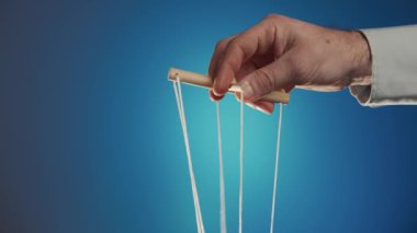 The hand of a man in a gray shirt controls a puppet using a wooden manipulator and strings on a blue background. Hand handling at puppet by pulling strings to make the character move. The concept of