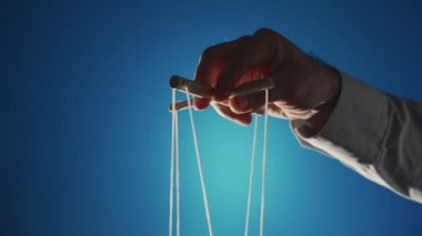 The hand of a man in a gray shirt controls a puppet using a wooden manipulator and strings on a blue background. Hand handling at puppet by pulling strings to make the character move. The concept of