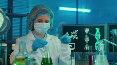 Dişi bir bilim adamı cam test tüpüne yeşil bir sıvı yerleştiriyor ve örneği inceliyor. Laboratuvarda beyaz önlüklü, mavi eldivenli ve maskeli bir kadın test tüpleri, mataraları ve bilgisayarı olan bir kadın.
