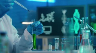 A female scientist pipettes a red drop into a test tube with a green liquid and examines the sample. A woman in a white medical coat, blue gloves and a mask in a laboratory with vials, flasks and