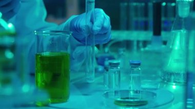 A woman scientist pipettes a green liquid into a test tube with white powder. The liquid bubbles up to form foam, a fizzy chemical reaction. Vaccine vials and glass flasks on the table. Biochemical