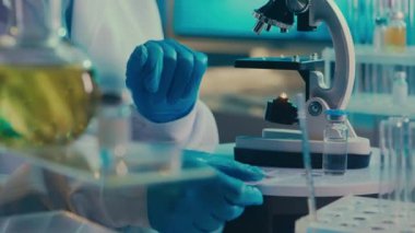 A female scientist applies a sample of red fluid with a glass pipette and examines it under a microscope. A woman in a white medical coat, blue gloves and a mask in a laboratory with test tubes