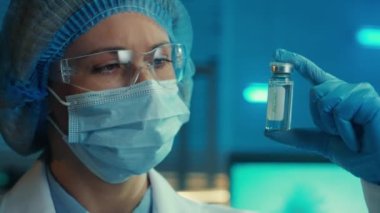 A woman examines a glass vial with a vaccine. Portrait of a female doctor in a goggles, white coat, medical mask, bonnet and blue gloves on the background of a laboratory or hospital. The concept of