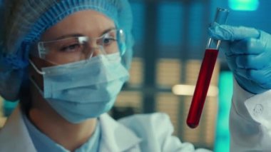 A woman carefully examines the red liquid in a test tube. Female doctor or researcher in goggles, bonnet, mask, white gown and blue gloves examines a sample. Biochemical laboratory, hospital. The