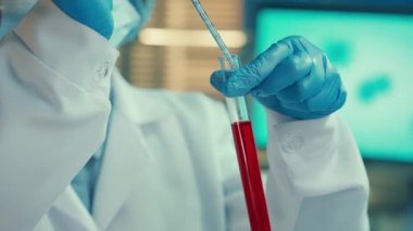 A woman fills a glass pipette with red liquid from a test tube and examines it carefully. Female doctor or researcher in goggles, bonnet, mask, white gown and blue gloves in biochemical laboratory or