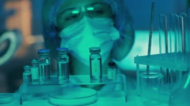 A woman doctor examines glass vials, flasks and test tubes. Female researcher or laboratory assistant in goggles, mask and gloves in a laboratory or hospital with blue light. The concept of medicine