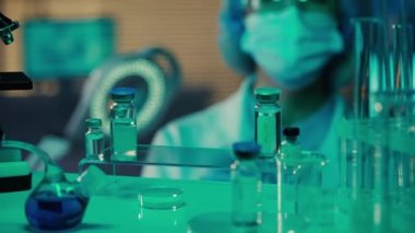 Kadın doktor cam şişeleri, mataraları, test tüplerini inceler ve verileri tablete koyar. Gözlüklü, maskeli ve eldivenli kadın araştırmacı labaratuvarda ya da hastanede mavi ışık altında. Bir raftan görüntüle