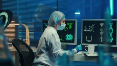 A doctor or scientist finishes work, takes off her mask, relaxes and drinks coffee from a cup. A woman in a white gown, gloves and bonnet sits near computer monitors. Biochemical laboratory with blue