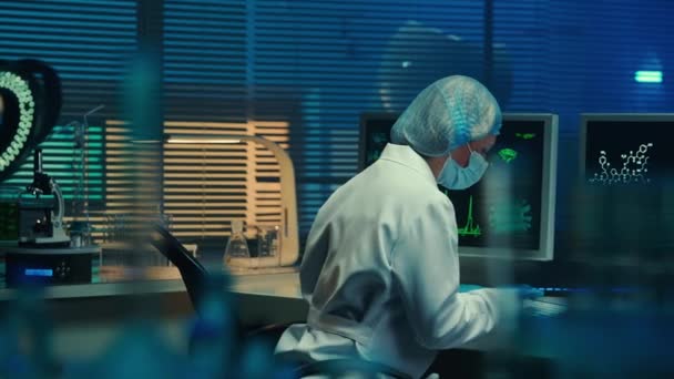 女性はコンピュータのキーボードを入力し モニター画面を見ています 白いコート マスク 手袋の科学者は 生化学研究所や病院に青い光で座っています 棚からの眺め — ストック動画