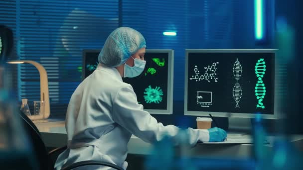 医生或科学家完成工作 摘下她的面具 从纸杯中放松下来并喝咖啡 一个穿着白色长袍 手套和帽子的女人坐在计算机监视器旁边 生物化学实验室 — 图库视频影像