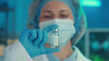 A woman examines a glass vial with a vaccine. Portrait of a female doctor in a white coat, medical mask, cap and blue gloves on the background of a laboratory or hospital. The concept of medicine