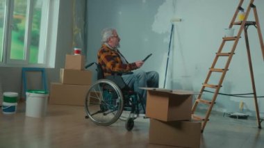 An elderly disabled man moves in a wheelchair plans renovation using a digital tablet. A pensioner checks building materials. Room with window, ladder, cardboard boxes, wallpaper rolls, paint bucket