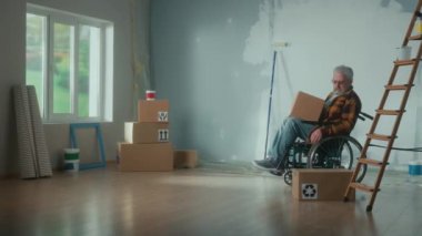 An elderly disabled man moves in a wheelchair with a cardboard box. The pensioner puts the box looks around and plans repairs. Room with window, stepladder, wallpaper rolls, roller and paint. Concept