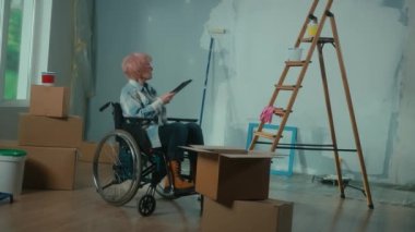 An elderly disabled woman moves in a wheelchair plans renovation using a digital tablet. A granny checks building materials. Room with window, ladder, cardboard boxes, wallpaper rolls, paint bucket