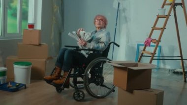 An elderly disabled woman moves in a wheelchair looks through the palette with colors. A granny plans repairs and chooses a paint color. Room with window, ladder, cardboard boxes, wallpaper rolls