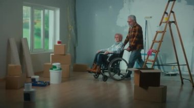 An elderly man brings an older woman in a wheelchair into a room. An elderly family couple plans repairs against the background of a window, ladder, cardboard boxes, buckets of paint, rollers and