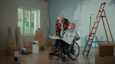 An elderly man and woman in a wheelchair look through a sheet with an apartment project and communicate. An elderly couple is planning repairs in the house against the background of a window