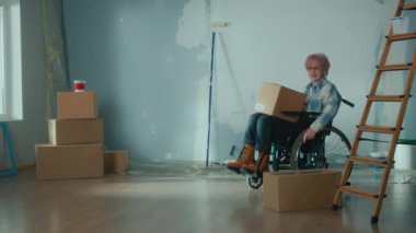 An elderly disabled woman moves in a wheelchair with a cardboard box. The granny puts the box looks around and plans repairs. Room with window, stepladder, wallpaper rolls, roller and paint. Concept