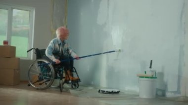 An elderly woman in a wheelchair paints a wall with white paint using a long roller. A disabled woman in the age makes repairs in the room. The concept of repair in the apartment performed by people