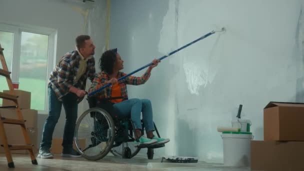 車椅子のアフリカ系アメリカ人女性が長いローラーを使って白いペンキで壁を塗ります 黒人女性と白人男性が部屋で修理をして喜んで笑う 若い夫婦だ — ストック動画