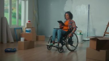 Young African American woman in a wheelchair is planning a renovation using a digital tablet. Disabled woman browses interior design ideas on the internet. Handicapped person on the background of