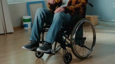 An elderly disabled man in a wheelchair. The disabilities male pensioner looks around and plans repairs. Handicapped person on the background of cardboard boxes, window and buckets of paint. The