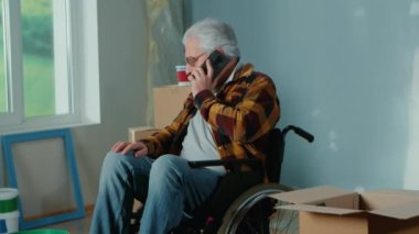 An elderly disabled man moves in a wheelchair and talks on a mobile phone. A pensioner takes a roller out of a cardboard box and plans repairs. Handicapped person on the background of cardboard boxes