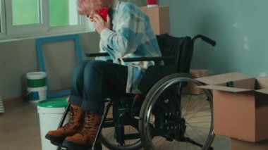An elderly disabled woman moves in a wheelchair and talks on a mobile phone. Granny inspects a bucket of paint and pulls a roller out of a cardboard box. Handicapped person on the background of window