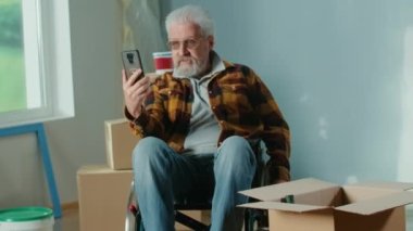 An elderly disabled man in a wheelchair talks on a video call using a mobile phone. A pensioner shows a wall and a window and plans repairs. Handicapped person on the background of cardboard boxes