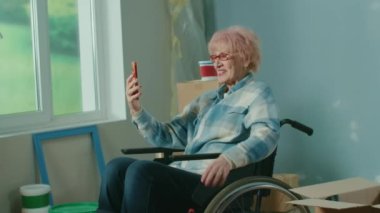 An elderly disabled woman in a wheelchair talks on a video call using a mobile phone. A granny shows a wall and plans repairs. Handicapped person on the background of cardboard boxes, window and