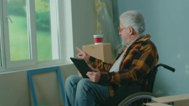 An elderly disabled man in a wheelchair plans renovation using a digital tablet. A pensioner checks building materials. Handicapped person on the background of window, cardboard boxes, paint buckets