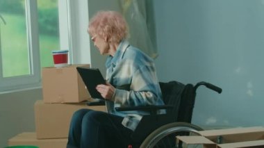 An elderly disabled woman in a wheelchair plans renovation using a digital tablet. A granny checks building materials. Handicapped person on the background of window, cardboard boxes, paint buckets