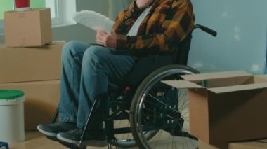 An elderly disabled man in a wheelchair looks through the palette with colors. A pensioner plans repairs and chooses a paint color. Handicapped person on the background of window, cardboard boxes
