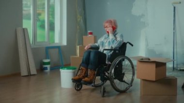 An elderly disabled woman moves in a wheelchair and talks on a mobile phone. Granny inspects a bucket of paint and pulls a roller out of a cardboard box. Room with window, ladder, wallpaper rolls. The