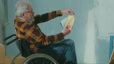 An elderly disabled man in a wheelchair looks through the palette with colors. A pensioner plans repairs and chooses a paint color. Handicapped person on the background of window, cardboard boxes