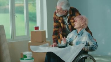 An elderly man and woman in a wheelchair look through a sheet with an apartment project and communicate. An elderly couple is planning repairs in the house against the background of a window