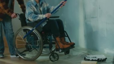 An elderly couple are doing repairs in the room. An aged woman in a wheelchair paints a wall with a long roller. An elderly couple smiles happily. The concept of renovation in the apartment
