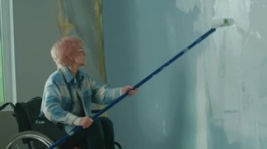 An elderly woman in a wheelchair paints a wall with white paint using a long roller. A disabled woman in the age makes repairs in the room. The concept of repair in the apartment performed by people