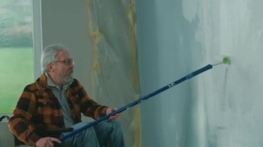 An elderly man in a wheelchair paints a wall with white paint using a long roller. A disabled man in the age makes repairs in the room. The concept of repair in the apartment performed by people with