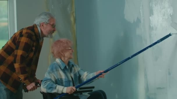 老人夫婦が部屋で修理をしている 車椅子の老女が長いローラーで壁を描く お年寄りのカップルは嬉しそうに微笑む アパートの改装の概念 — ストック動画