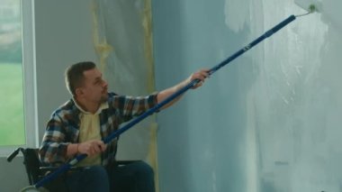 A young man in a wheelchair paints a wall with white paint using a long roller. A disabled man makes repairs in the room against the background of cardboard boxes and buckets of paint. The concept of