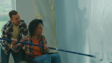 An African American woman in a wheelchair paints a wall with white paint using a long roller. A black woman and a white man make repairs in the room and laugh happily. A young couple. The concept of