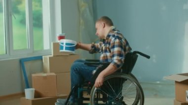 A young man in a wheelchair is talking on a mobile phone. The disabled person takes a bucket of paint and a roller and sharing plans to repairs a room. Handicapped person on the background of