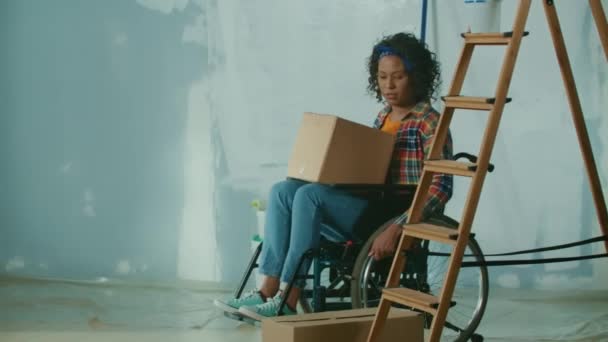 車椅子に乗ったアフリカ系アメリカ人の若い女性が 膝の上に段ボール箱を持った部屋に入ってきた 身障者の女性は部屋の修理を見 計画する 身障者用 — ストック動画