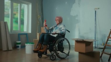 An elderly disabled woman in a wheelchair talks on a video call using a mobile phone. A granny shows a wall and plans repairs. A room with a window, ladder, wallpaper, cardboard boxes and a bucket of