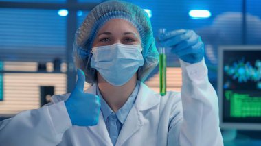 A woman scientist holds a test tube with a green liquid in her hand and shows a thumbs up. A woman in blue gloves, a white medical gown and a mask shows a recommending gesture, like. Close up portrait