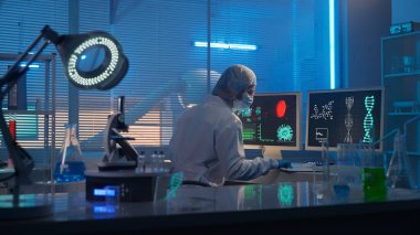 Tıbbi gelişim laboratuvarı. Kadın bir bilim adamı bilgisayarda çalışıyor ve analiz verilerini kontrol ediyor. Modern bir laboratuvarın arka planına karşı deney tüpleri, mataraları ve...